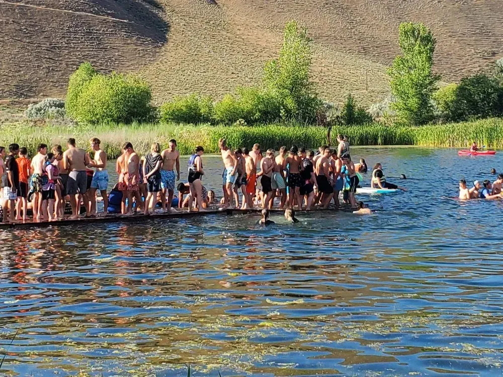 Crowd standing on a dock at a lake, with swimmers in blue water and hills in the background