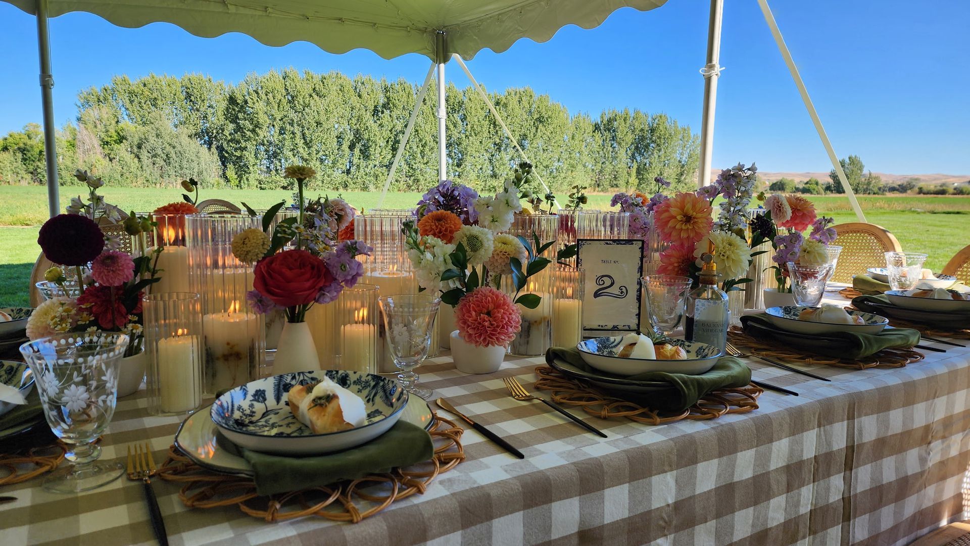 Outdoor buffet table under a tent, set with flowers, candles, and plates on a checkered cloth.