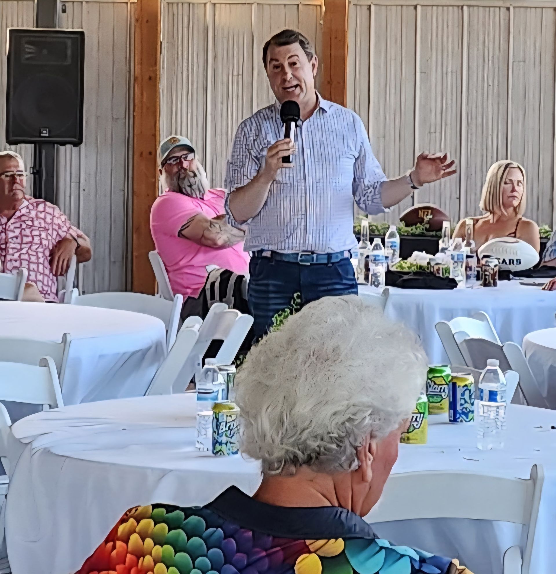 Man speaking into a microphone at a casual indoor gathering with seated guests and white tablecloths