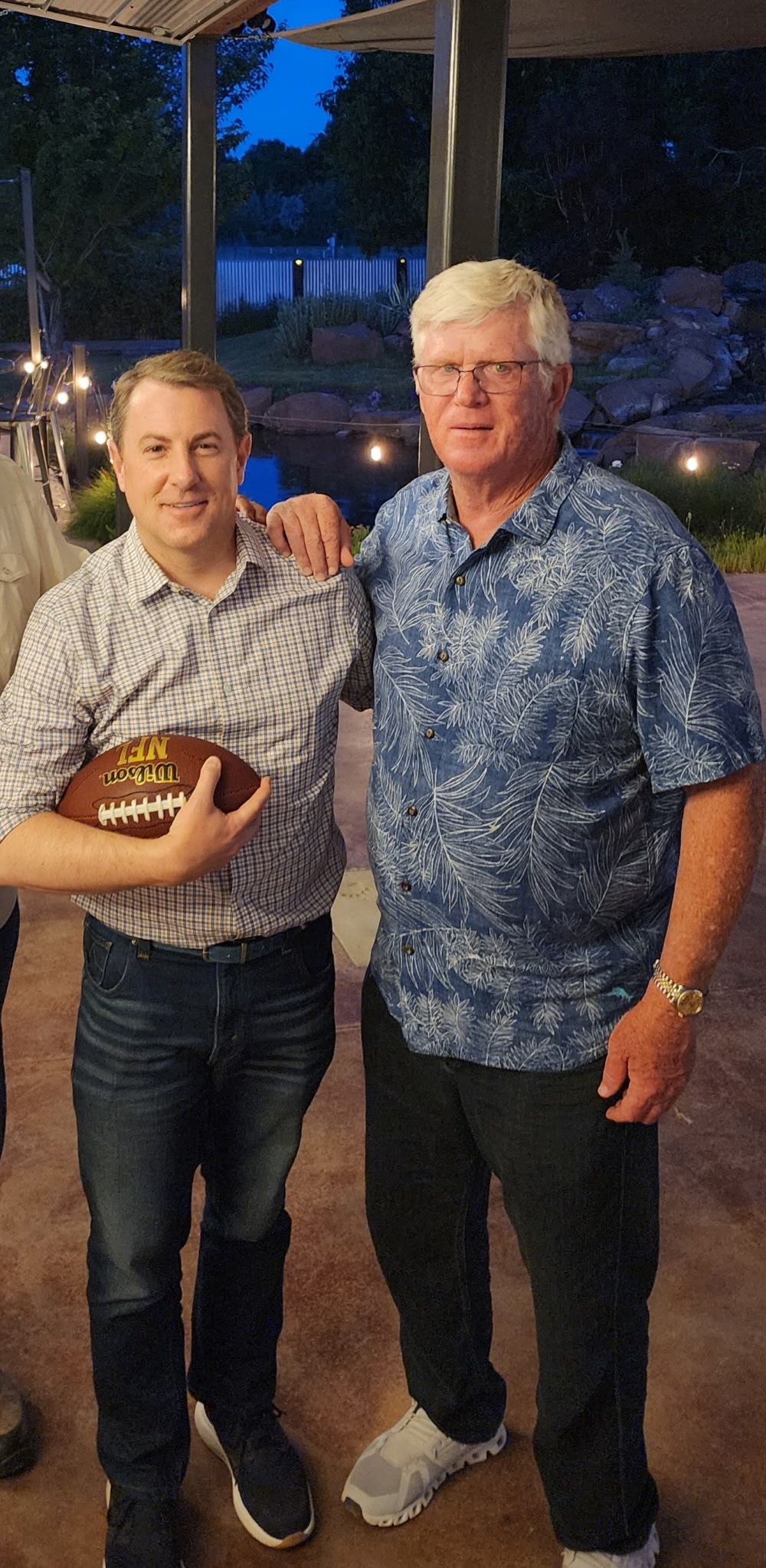 Two men standing outdoors at night, holding a football, with trees and lights in the background.