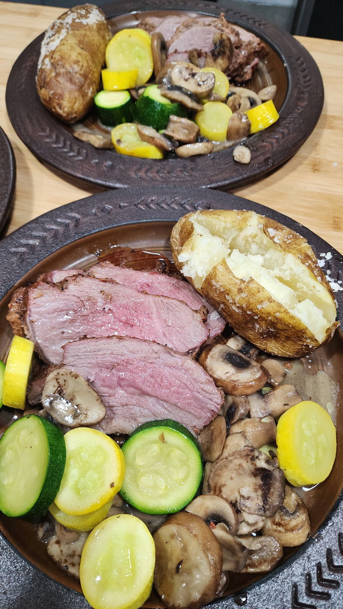 Two plates of steak with baked potato, mushrooms, and sliced zucchini on a wooden table