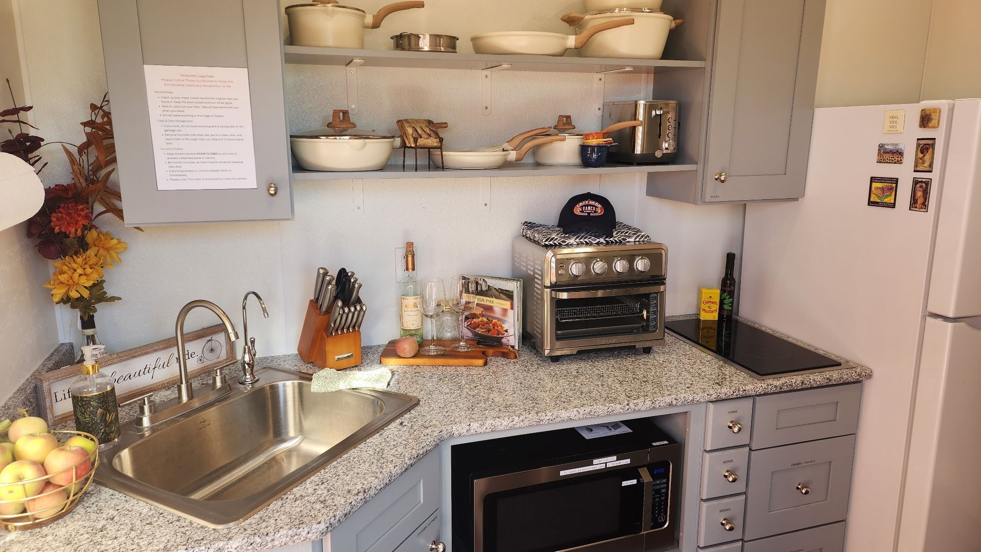 Compact kitchen with stainless sink, speckled countertops, open shelves, toaster oven, and white fridge