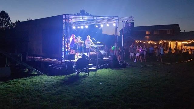 Outdoor concert at dusk with a lit stage, musicians performing, and a crowd gathered on the grass