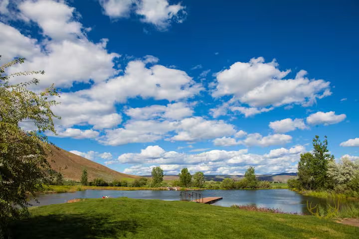 Blue lake with a small dock, green grass, trees, and a cloudy sky over hills