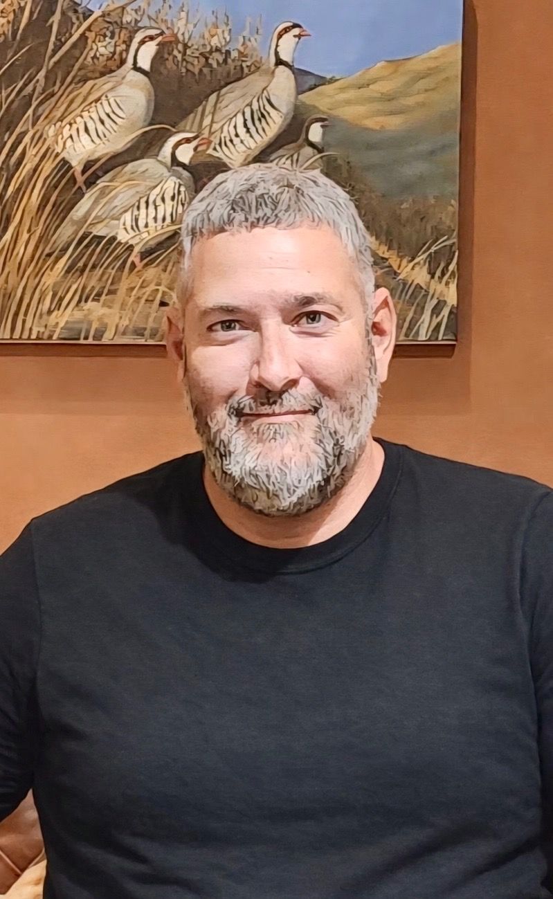 Man in black shirt smiling indoors, standing in front of a framed bird painting.