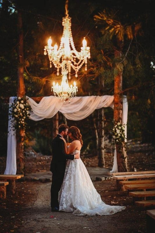 Couple embracing beneath chandeliers at an outdoor evening wedding ceremony
