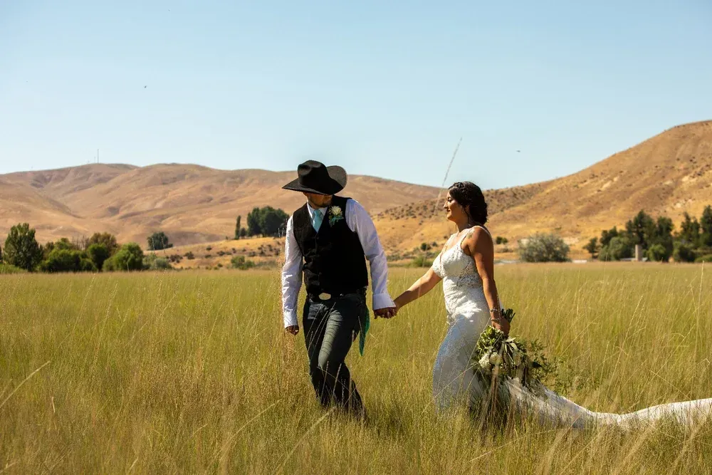 Couple holding hands and walking through a grassy field with hills in the background