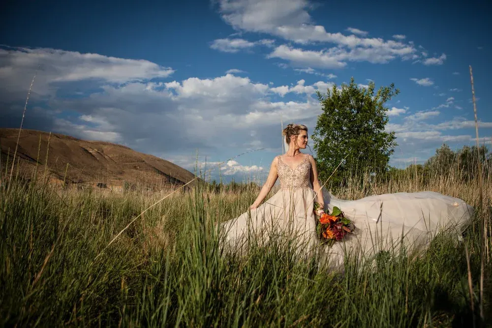 Bride in a white gown standing in tall grass under a blue sky, holding a colorful bouquet.