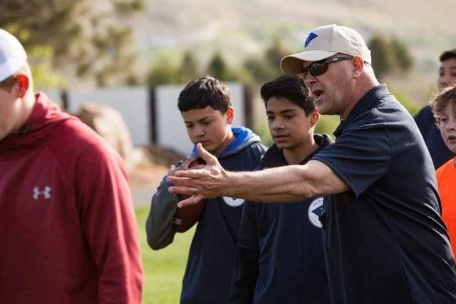 Coach in navy cap gestures to players during outdoor football practice.