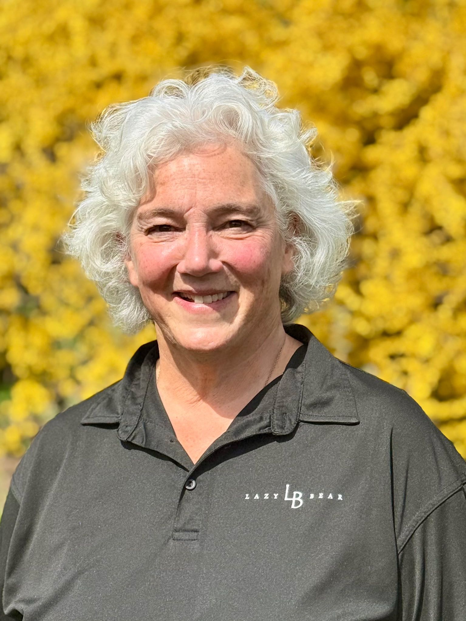 Smiling person with curly white hair in a dark polo, standing before bright yellow flowers
