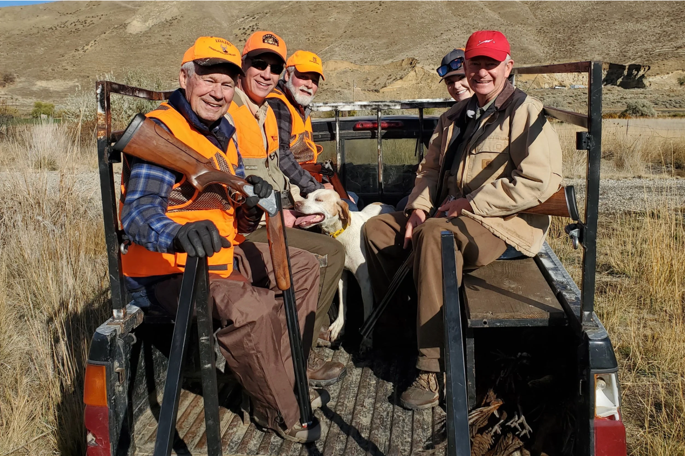 Four hunters in orange vests and hats pose with a deer on an open trailer in a grassy field.