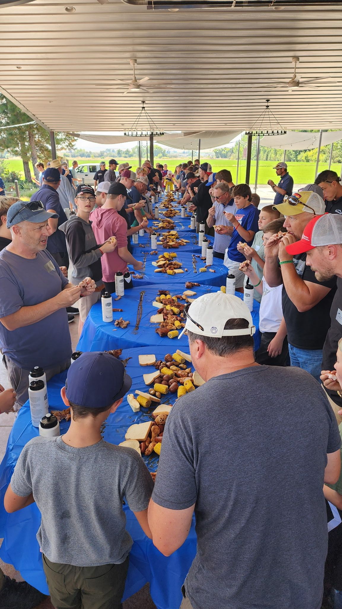 Long picnic tables under a pavilion, crowded with people sharing a meal on blue tablecloths.