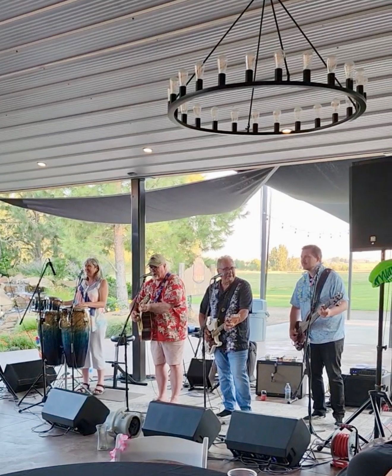 Live band performing under a covered outdoor pavilion, with microphones, guitars, and percussion onstage.