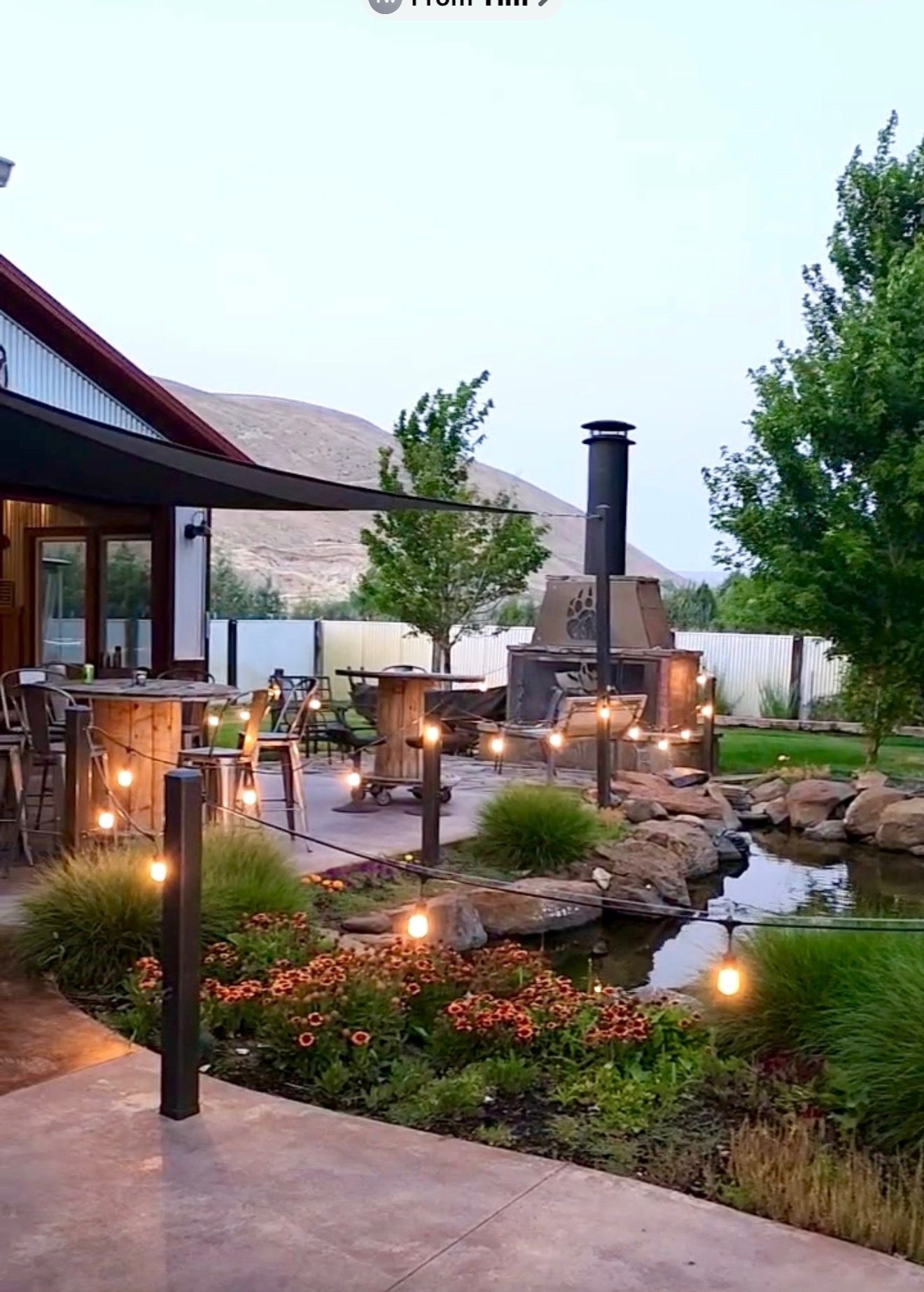 Outdoor patio with stone fire pit, string lights, and lush garden landscaping at dusk
