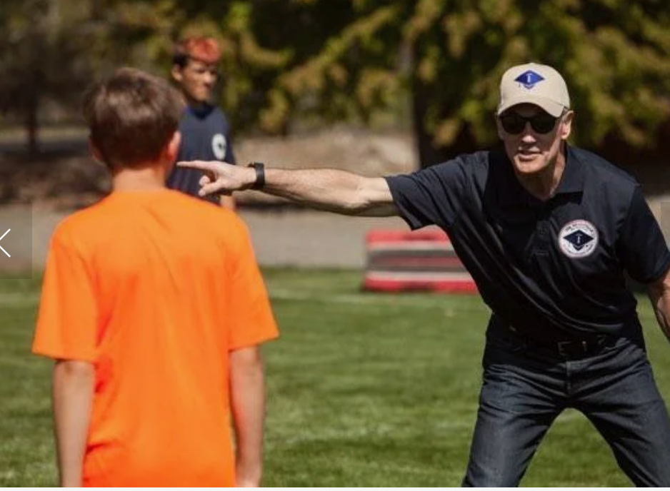 Coach in black shirt and cap points on a field to a person in an orange shirt.