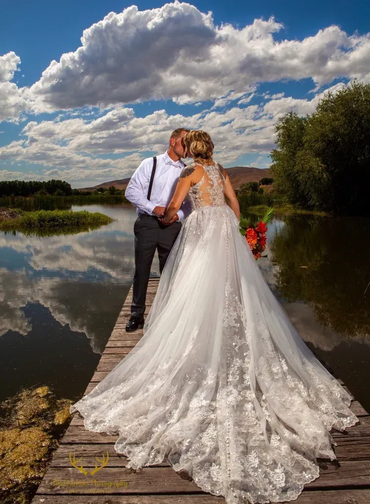 Bride and groom kissing on a wooden dock by a lake under a cloudy blue sky