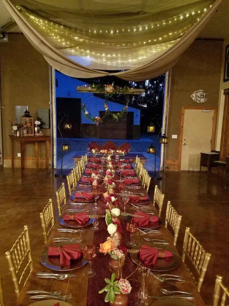 Long banquet table with burgundy place settings and gold chairs under draped lights in a decorated hall