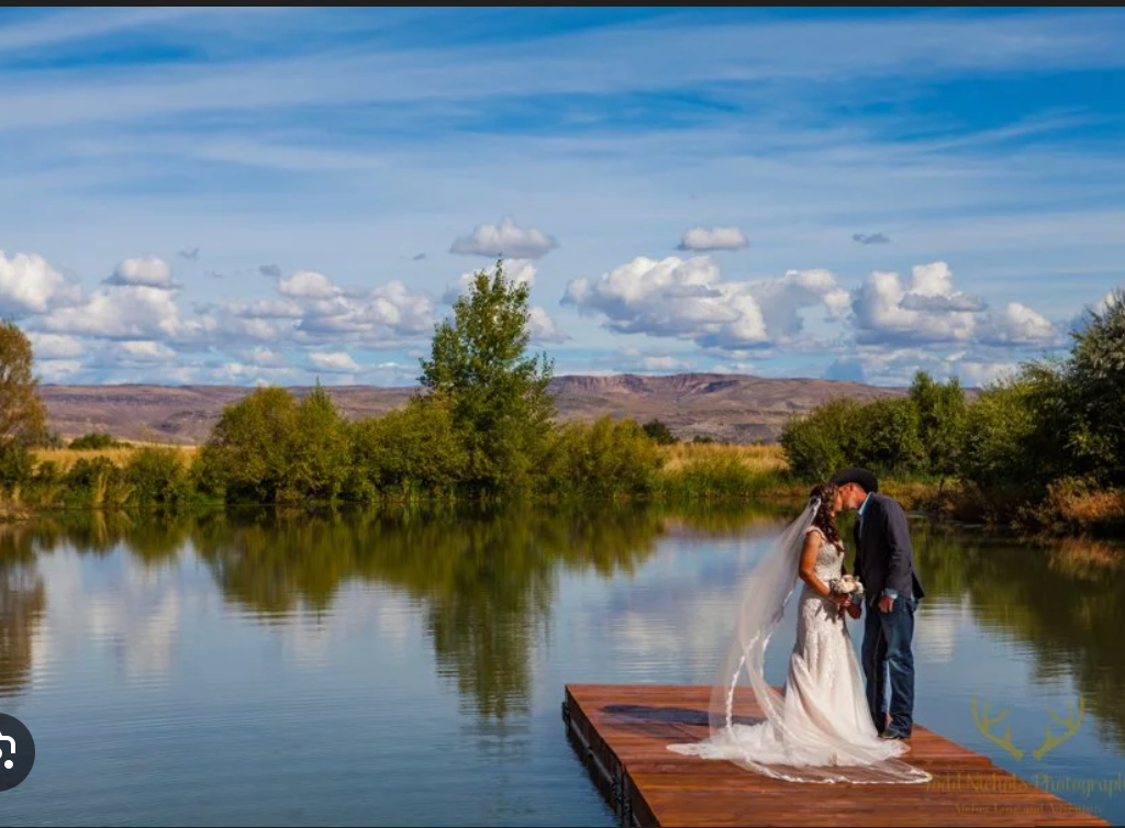 Bride and groom embrace on a dock by a calm lake under a blue sky with clouds