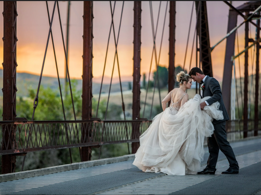 Bride and groom embrace on a bridge at sunset, with city lights and warm sky in the background.
