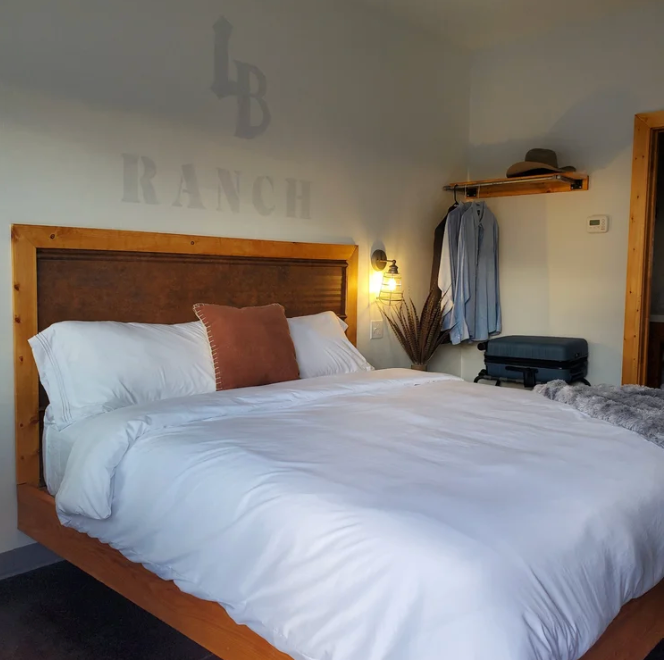 Cozy bedroom with white bedding, wooden headboard, wall lamp, and “RANCH” decor above the bed