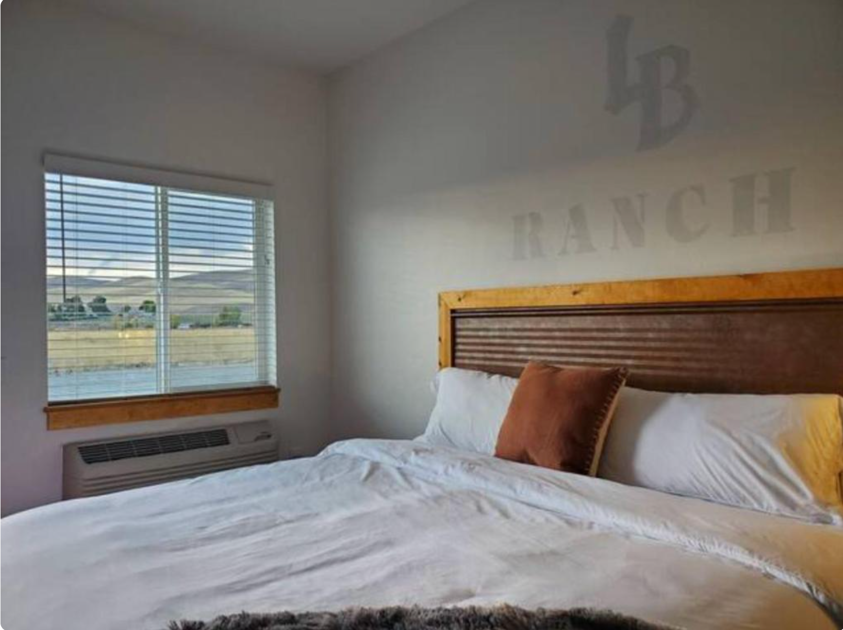 Bright bedroom with a wooden headboard, white bedding, and a window overlooking fields.