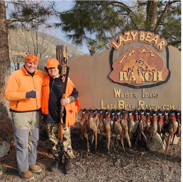 Two people in orange gear posing beside the Lazy Bear Ranch sign and hanging fish in Weiser, Idaho