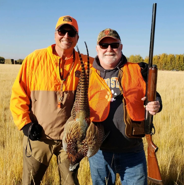 Two hunters in orange vests hold a pheasant and a shotgun in a field under clear sky