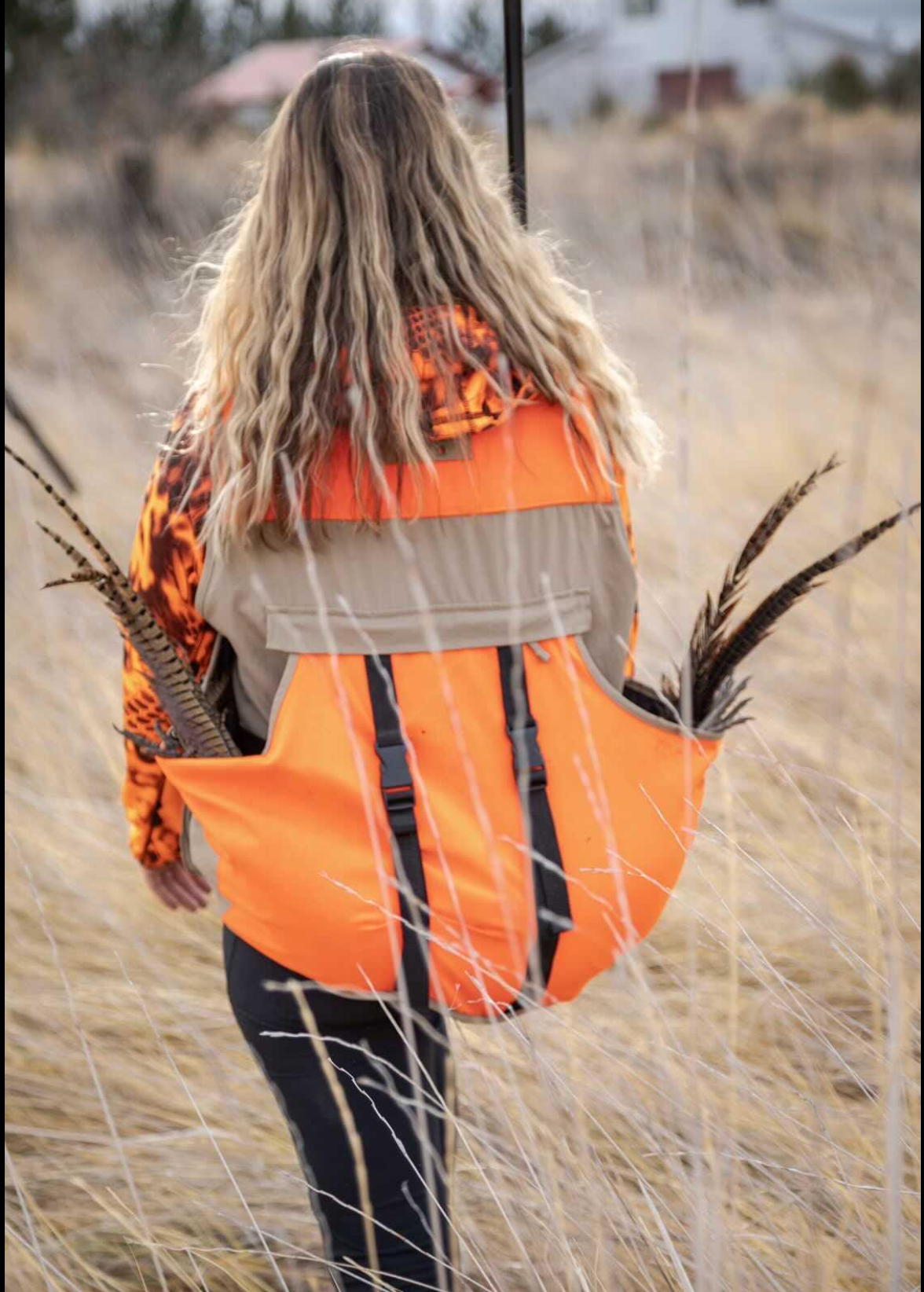 Person in an orange safety vest walking through tall dry grass outdoors, seen from behind.