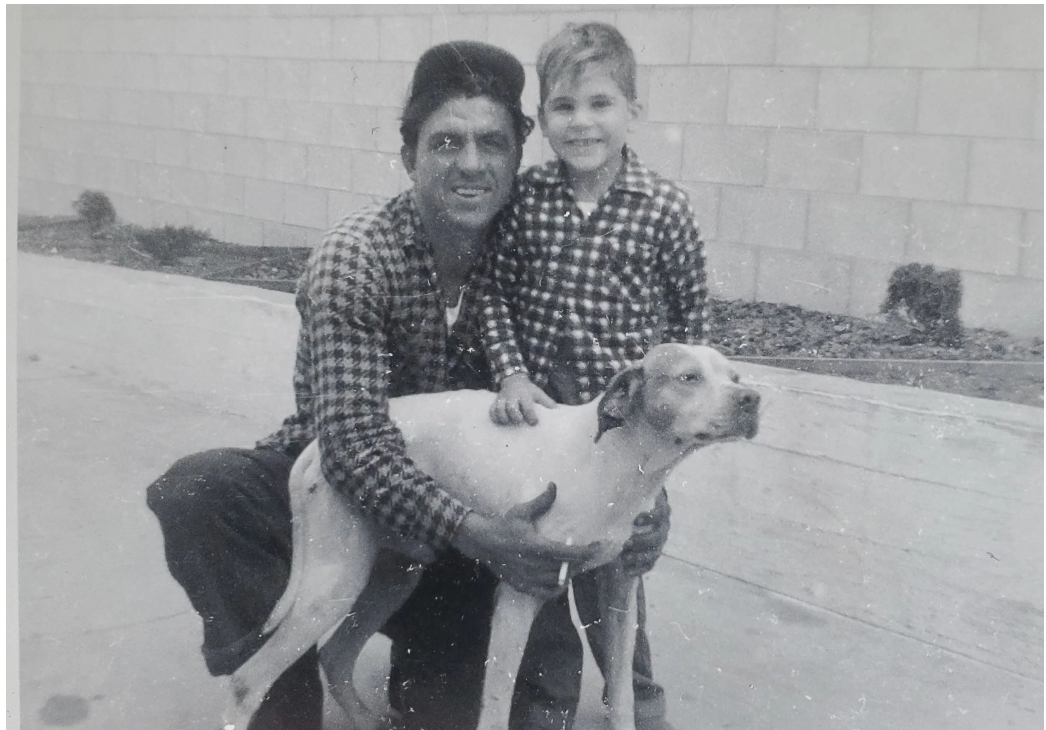 Two people in plaid shirts pose with a dog on a sidewalk beside a brick wall