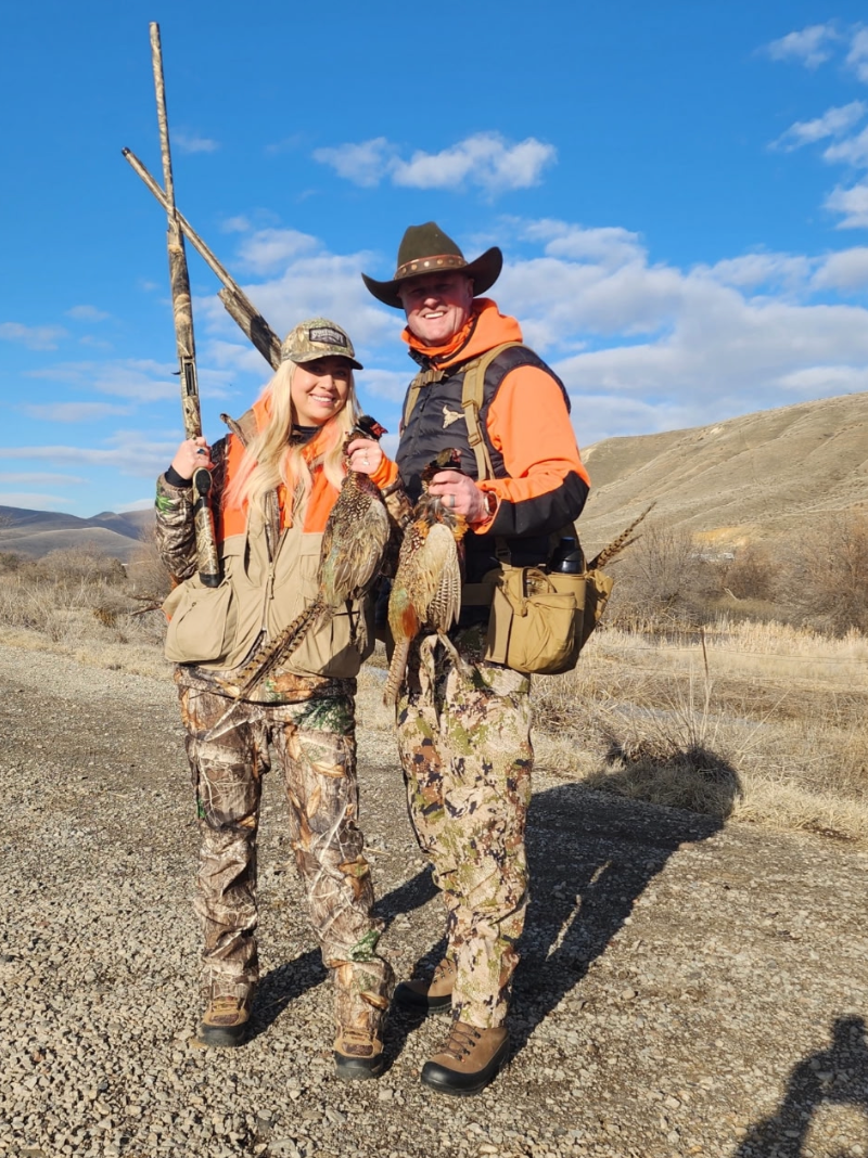 Two hunters in camo and orange vests holding rifles in a dry, open field under a blue sky
