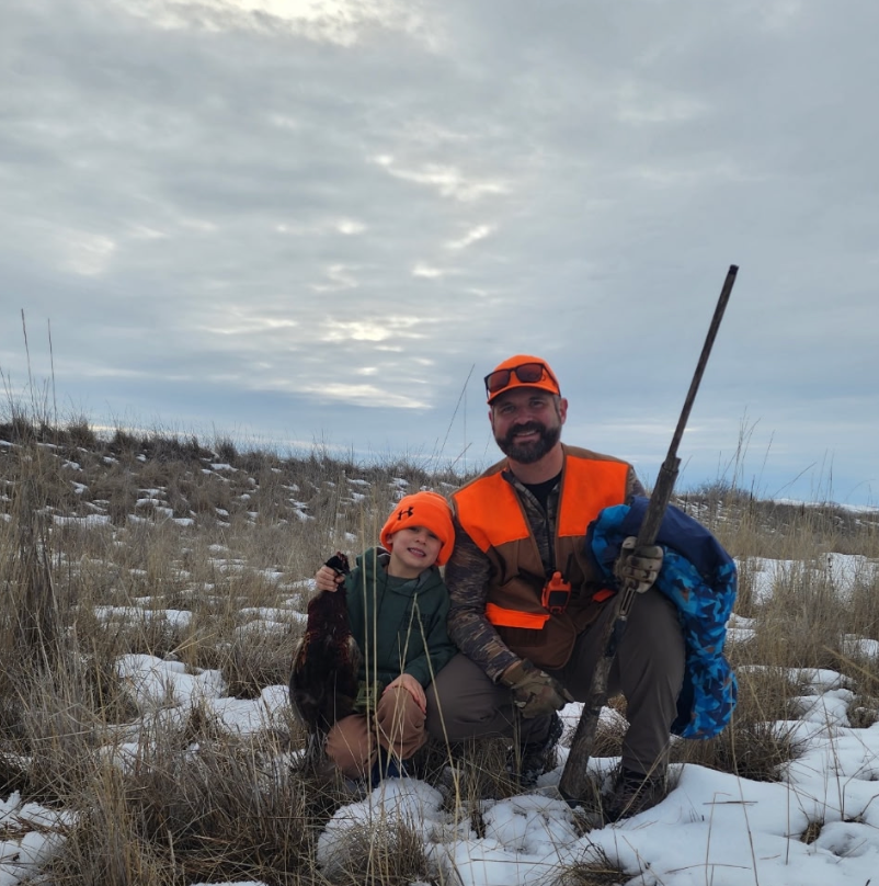 Hunter in orange gear kneels in snowy field with two dogs and a rifle at dusk