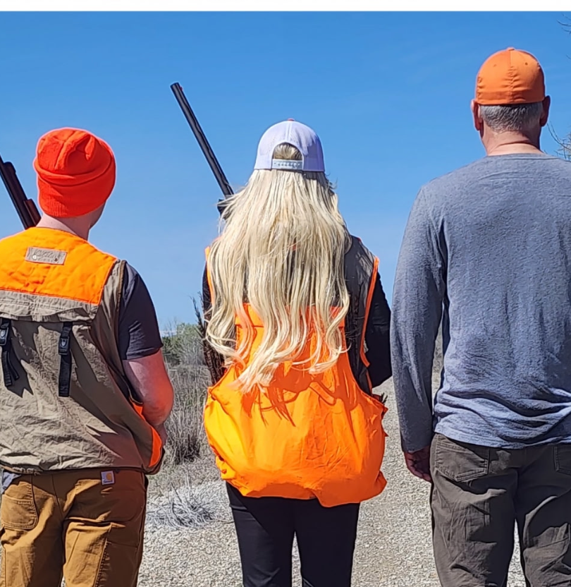 Three hikers seen from behind on a rocky trail under a blue sky, one carrying a rifle.