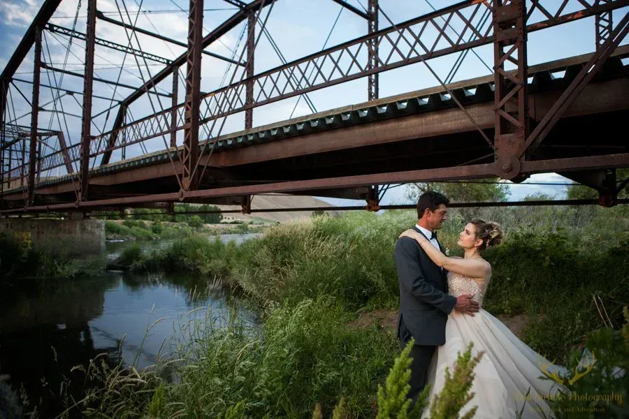 Couple in wedding attire embracing beside a river under a steel bridge