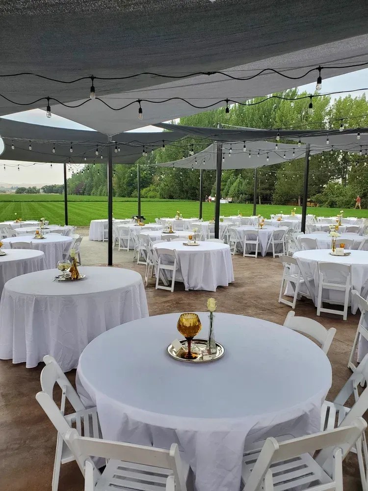 Outdoor wedding reception setup with round white tables, chairs, and gray umbrellas on a patio by a green lawn