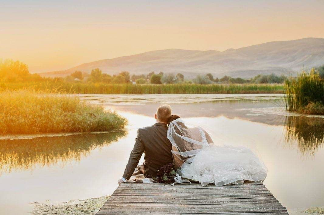 Couple sitting on a dock by a calm lake at sunrise, with a flowing white dress and warm golden light