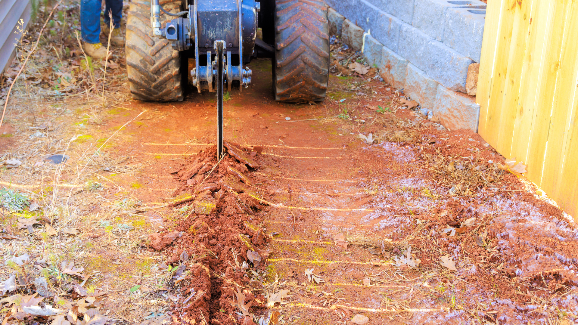 A worker in blue workwear stands beside a yellow industrial drill boring into the ground outdoors.
