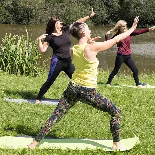 People practicing the archer pose on grass by a pond.