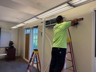 Person on a ladder adjusts a wall-mounted air conditioner in a hallway.