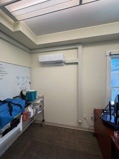 Small office room with whiteboard, wall-mounted AC, desk, and blue bags on a table