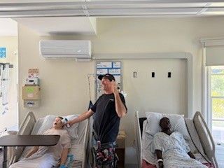 Hospital room with two patients in reclining chairs and a man standing between them holding a phone.