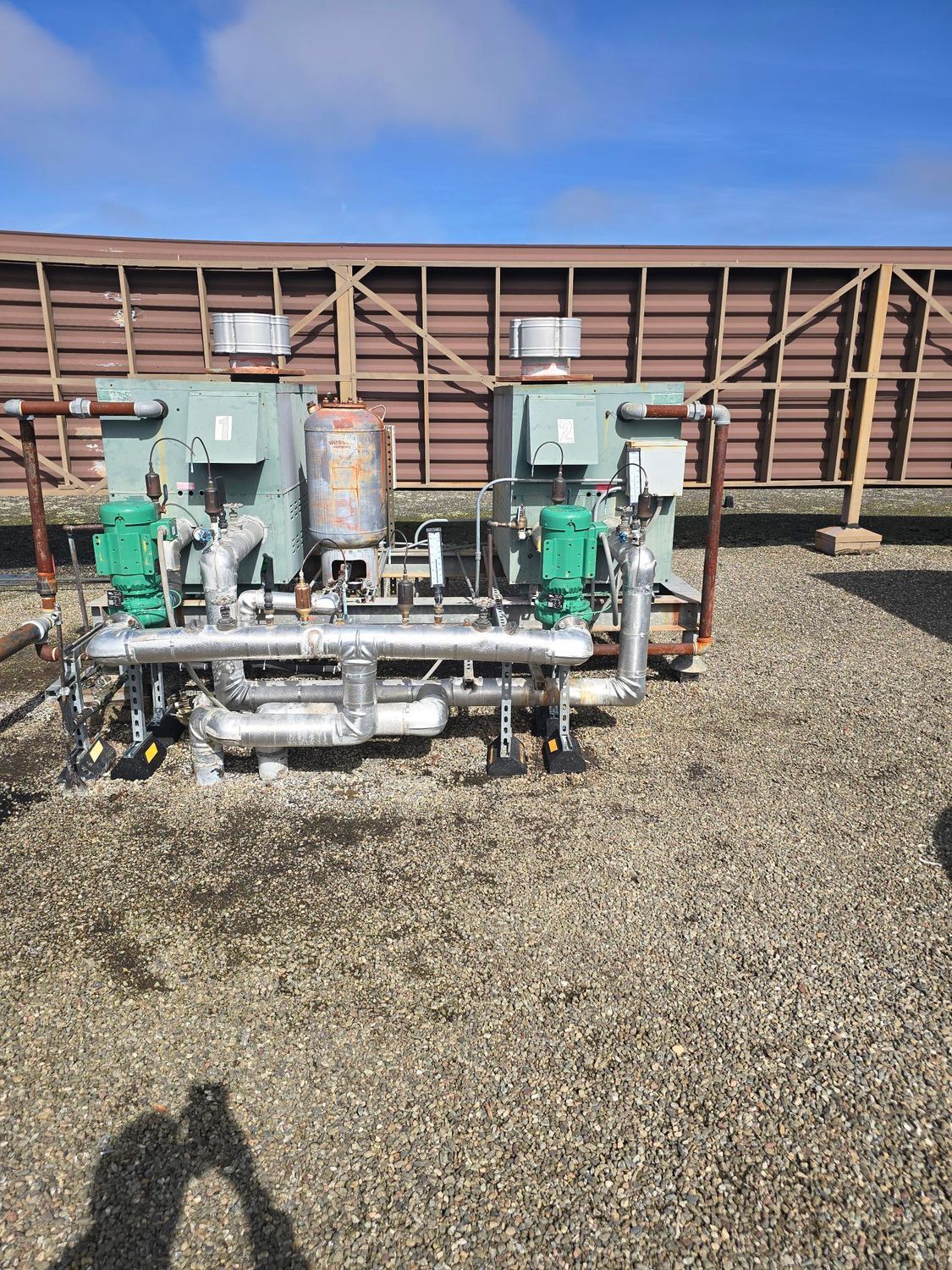 Industrial pump and pipe equipment on gravel beside a large brown structure under a blue sky
