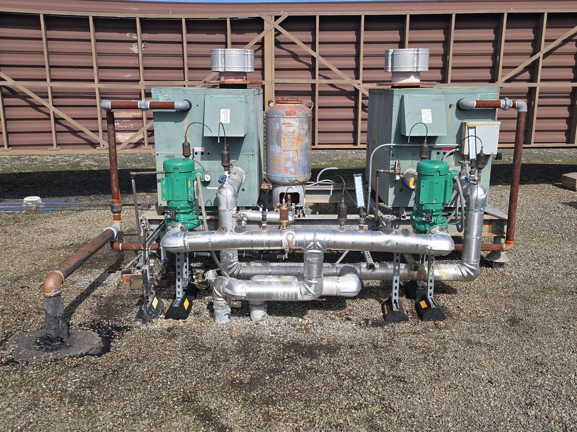 Two green industrial pumps and pipes on gravel in front of a wooden fence