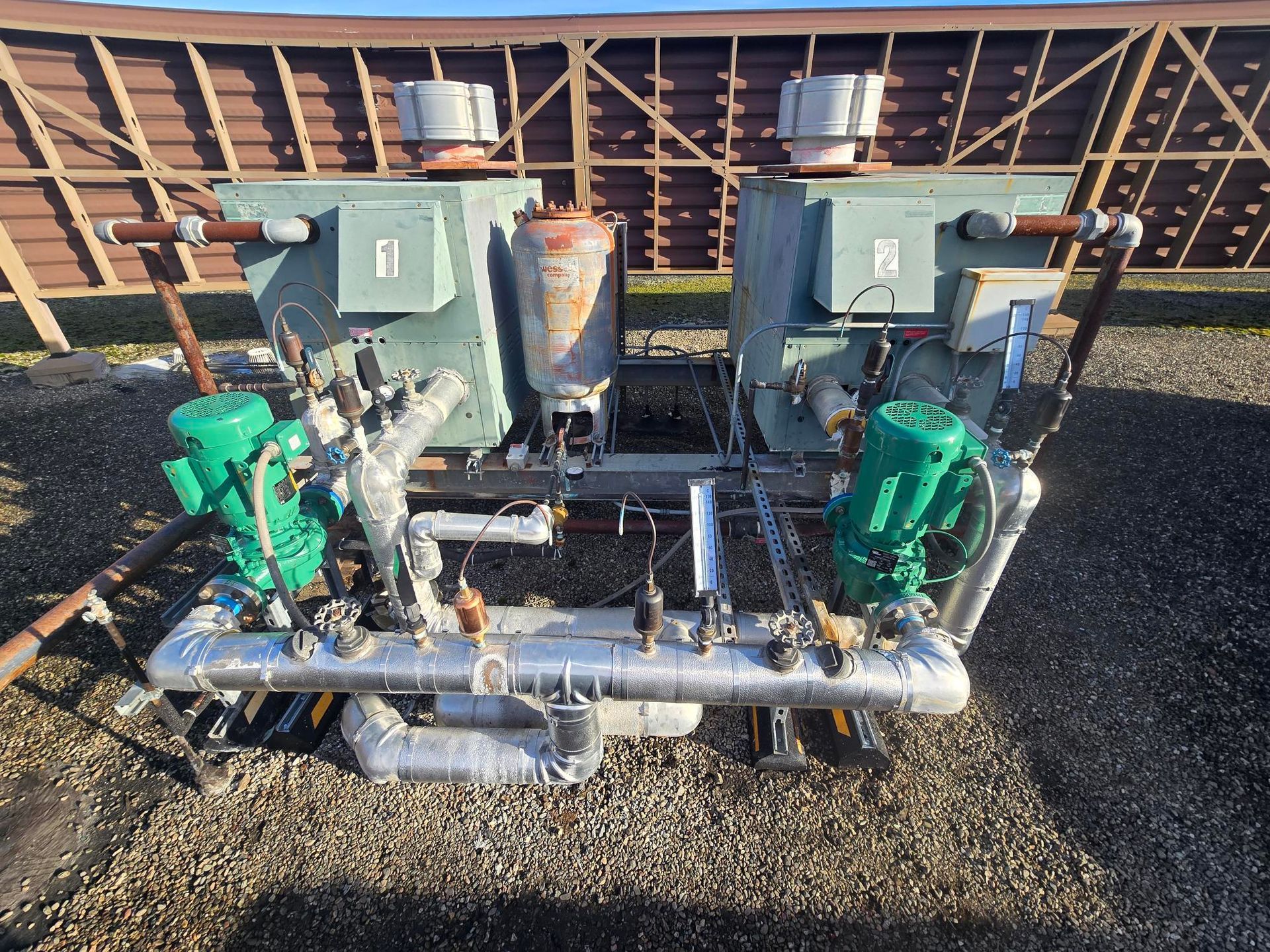 Outdoor mechanical equipment with green pumps and silver pipes mounted on a gravel surface.