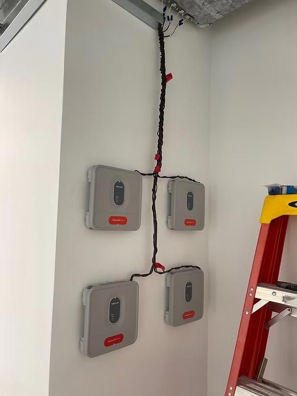 Four gray wall-mounted electrical boxes with red labels and exposed wiring near a red ladder in a utility room