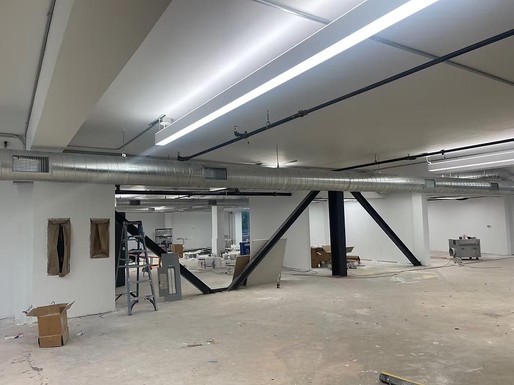 Empty industrial basement with exposed beams, pipes, and scattered construction materials under bright strip lights