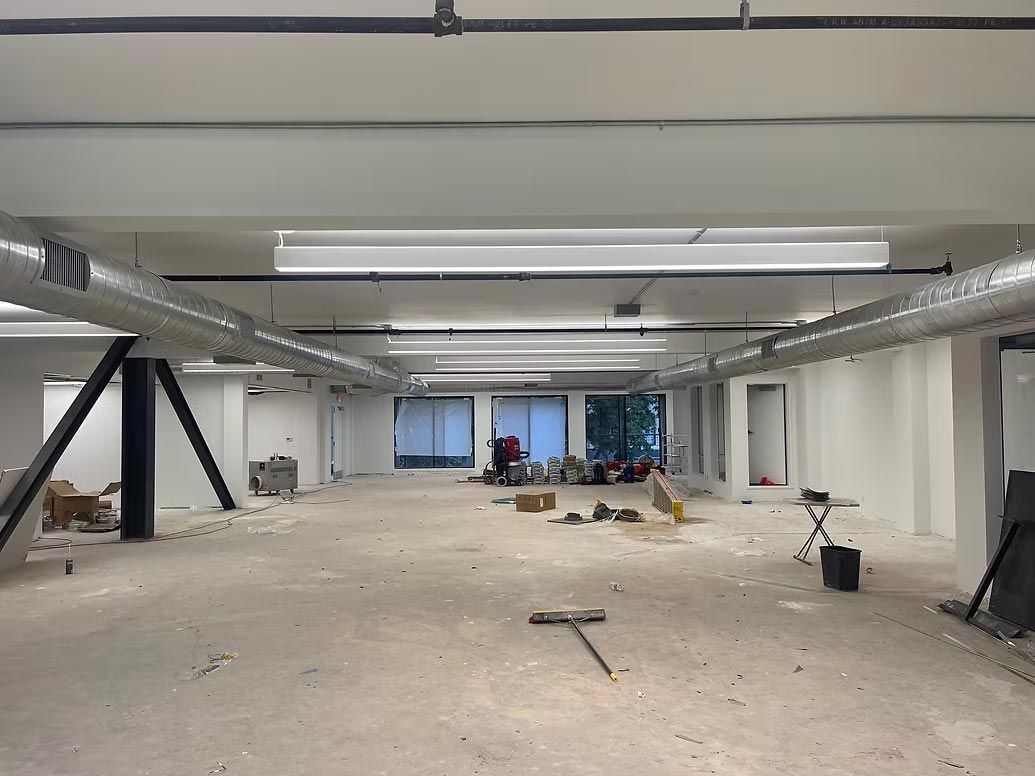 Empty unfinished office interior with exposed ceiling, scattered tools, and large windows at the far end