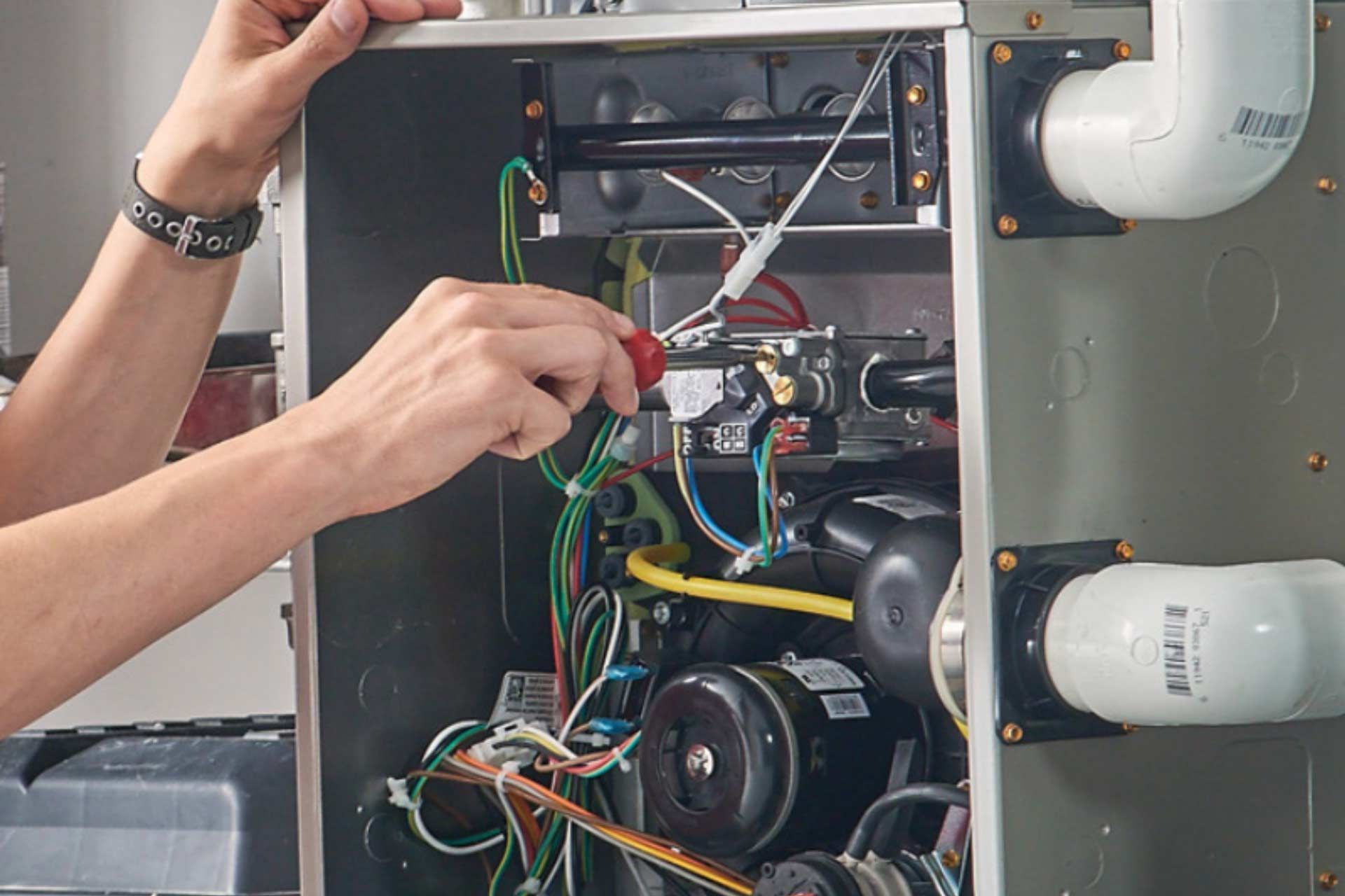 Technician wiring a furnace control panel with exposed wires and pipes nearby