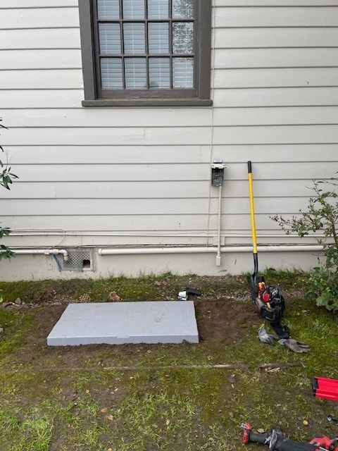Backyard wall with window, utility pipes, a gray board on the ground, and tools leaning beside it.