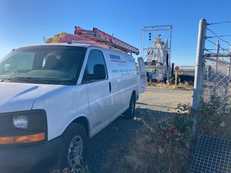 White utility van with ladder parked beside a fenced industrial site under a clear blue sky.