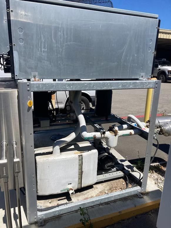 Outdoor industrial equipment unit in a metal frame with pipes, tanks, and hoses on a concrete lot